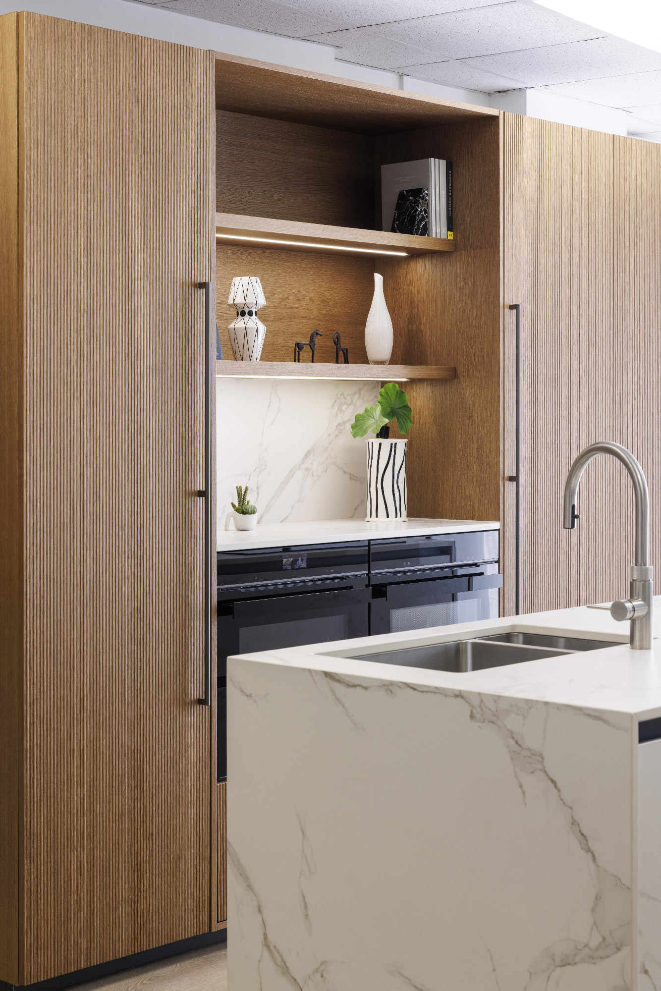 Cucina moderna con colonne in legno scanalato, isola in marmo bianco venato, doppio lavello inox e dettagli decorativi minimal — design contemporaneo showroom Zecchinon Londra