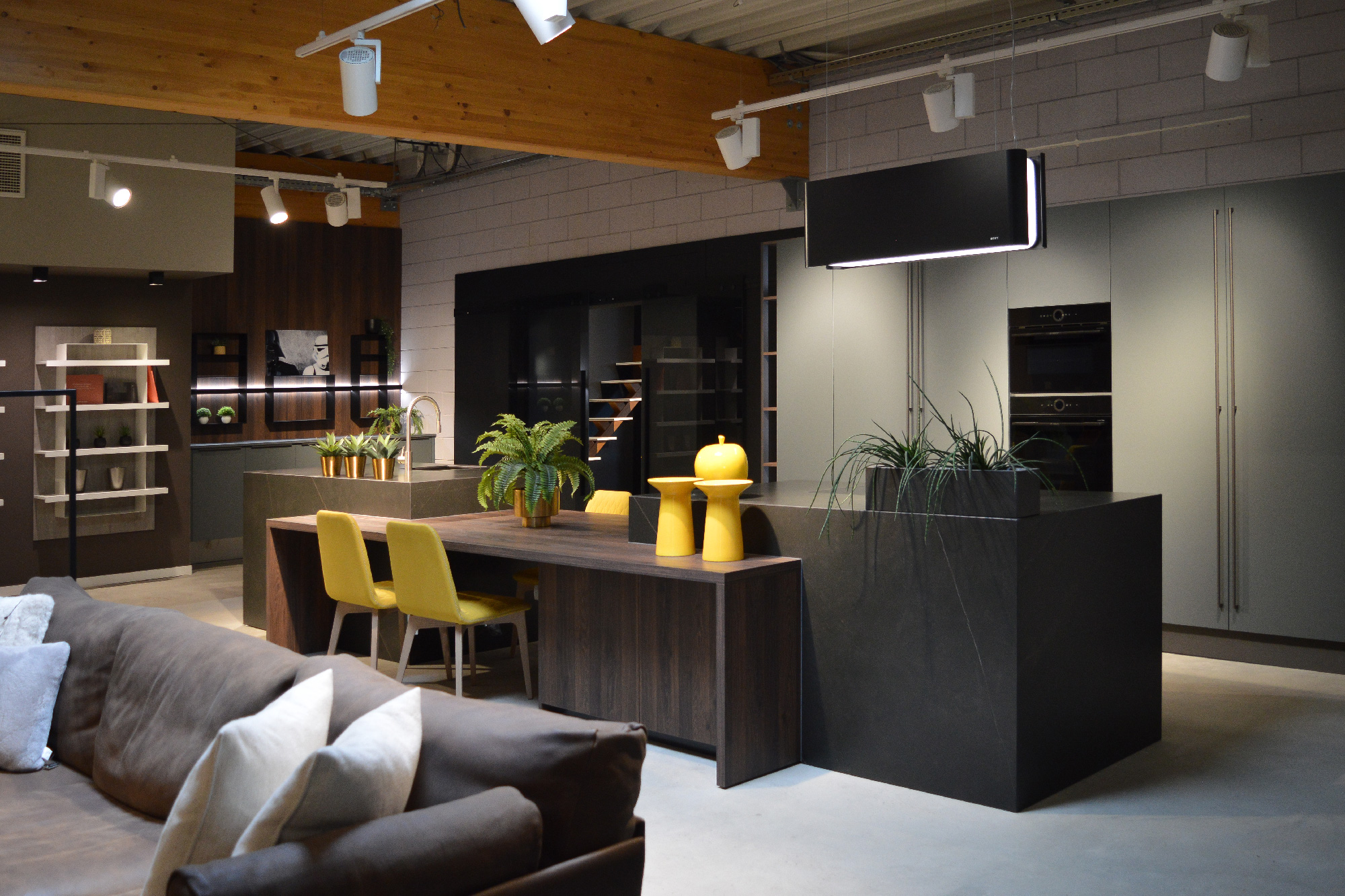 Showroom cucina moderna B&B Concept a Messancy con isola in marmo nero, sedie gialle, piante decorative e illuminazione a binario.