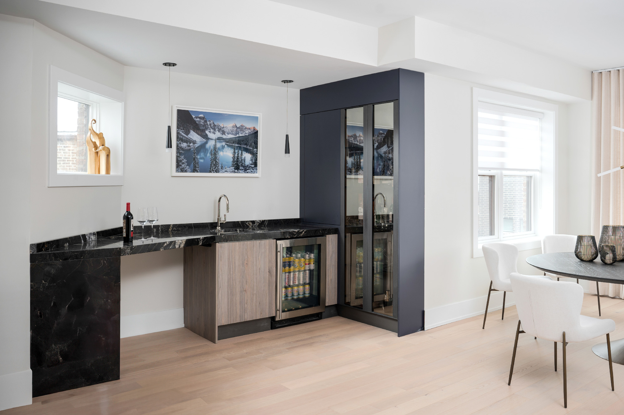 Zona bar raffinata con top in marmo nero, frigoriferi vetrati da incasso e quadro paesaggistico, accanto a zona pranzo con sedie bianche e tavolo rotondo. Progetto Zecchinon e Archisesto a Chicago.
