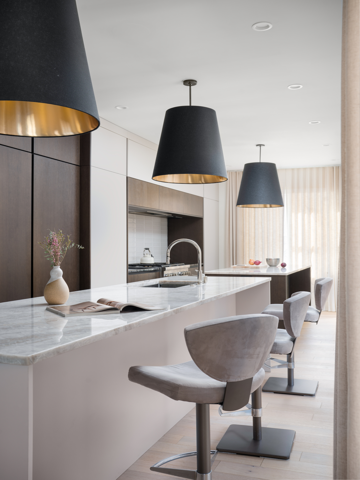 Dettaglio frontale di isola cucina con top in marmo, sedute imbottite e sfondo con pensili in legno scuro, backsplash bianco lucido e lampada nera con interno dorato. Progetto Zecchinon e Archisesto a Chicago.