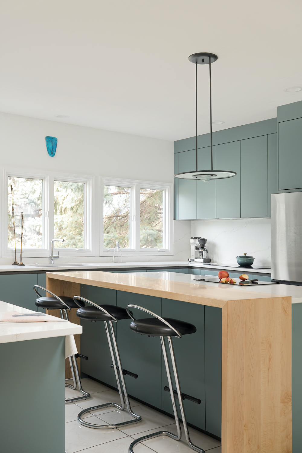 Isola cucina in laccato opaco e inserto in legno con piano a induzione, tre sgabelli con seduta in pelle nera e struttura in acciaio, illuminazione a sospensione moderna.